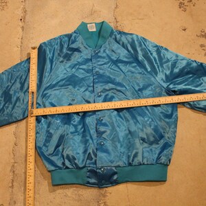 May include: Teal satin bomber jacket with a button-up front, teal ribbed collar, cuffs, and hem. The jacket is laid flat on a concrete surface. A wooden ruler is placed across the jacket to measure its length.