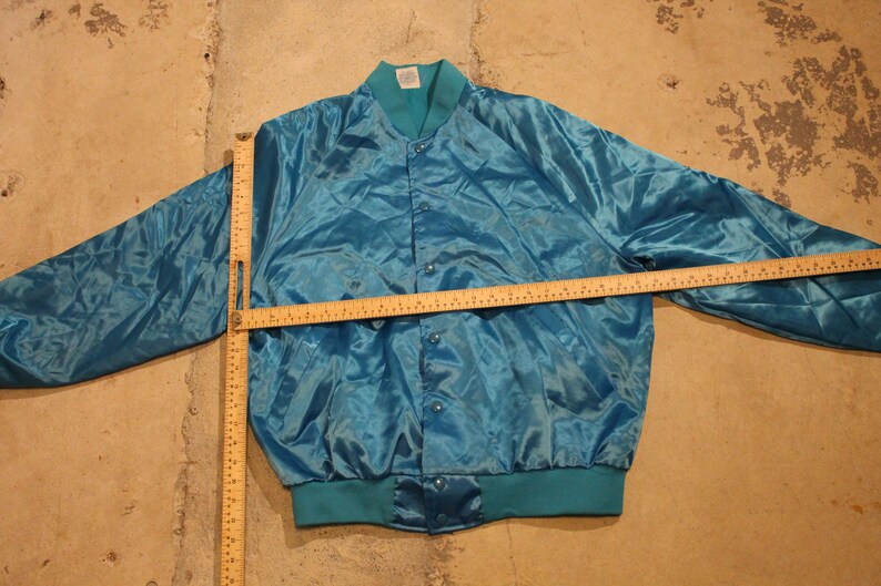 May include: Teal satin bomber jacket with a button-up front, teal ribbed collar, cuffs, and hem. The jacket is laid flat on a concrete surface. A wooden ruler is placed across the jacket to measure its length.