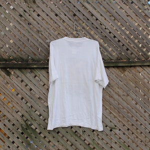Pode incluir: Uma camiseta branca lisa pendurada em uma cerca de madeira. A camiseta &eacute; lisa e n&atilde;o tem gr&aacute;ficos ou texto vis&iacute;vel.