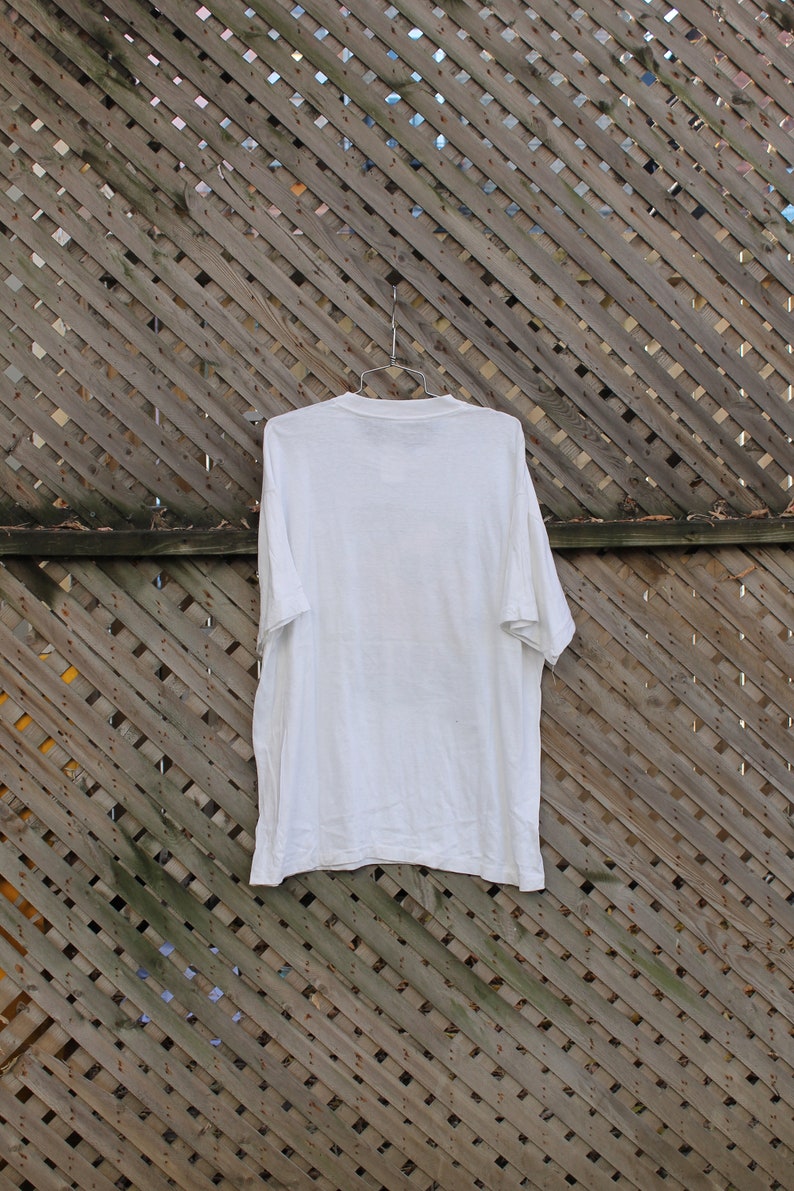 Pode incluir: Uma camiseta branca lisa pendurada em uma cerca de madeira. A camiseta &eacute; lisa e n&atilde;o tem gr&aacute;ficos ou texto vis&iacute;vel.