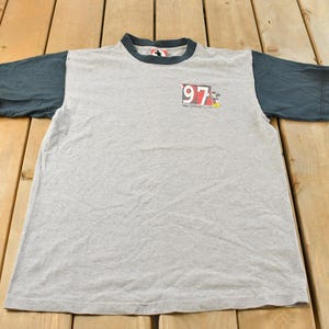 May include: A gray t-shirt with dark blue sleeves and collar. The shirt features a small graphic with the number "97" and a cartoon character. The shirt is made of a soft, likely cotton, material.