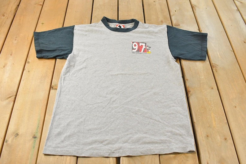 May include: A gray t-shirt with dark blue sleeves and collar. The shirt features a small graphic with the number "97" and a cartoon character. The shirt is made of a soft, likely cotton, material.