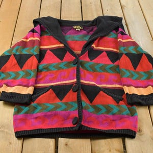 Puede incluir: Un cárdigan de manga larga con capucha que presenta un patrón geométrico vibrante en tonos rojo, negro, verde azulado, morado y naranja. La prenda de punto incluye una capucha negra, puños acanalados negros, una tapeta negra con tres botones grandes.