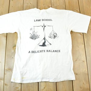May include: White t-shirt featuring a balance scale graphic, with the text "LAW SCHOOL" above and "A DELICATE BALANCE" below. The scale has two hanging trays with illustrations. The shirt is laid flat on a wooden surface.