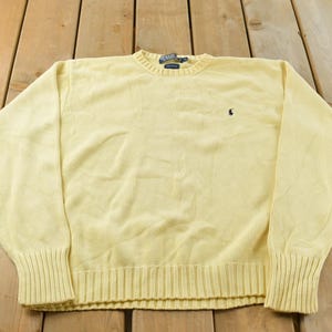 May include: A pale yellow long-sleeve crewneck sweater featuring a ribbed collar, cuffs, and hem. The garment displays a small, dark-colored embroidered logo on the left chest area and a brand label stitched inside the back of the neckline.