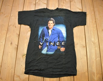 May include: Black t-shirt with a faded image of a man wearing a blue shirt and the word "VINCE" printed on the front.