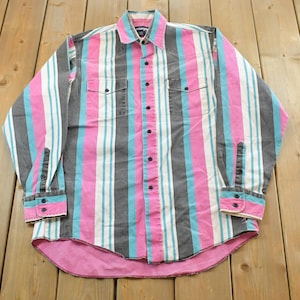 Puede incluir: Camisa abotonada con rayas verticales rosas, grises, blancas y turquesas. La camisa tiene dos bolsillos en el pecho, puños abotonados y un dobladillo rosa. El cuello es rosa con un pequeño logotipo.