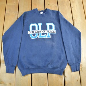 May include: A navy blue sweatshirt featuring the text "OLD" in white and light blue block letters, with "OUR LADY OF PEACE" above the letters. The sweatshirt is made of a soft material and has long sleeves.