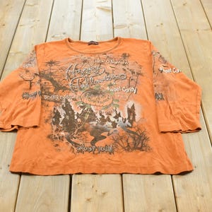 May include: An orange long-sleeve shirt featuring a Halloween-themed graphic print. The design includes a jack-o'-lantern, a castle, and bare trees, with text reading Happy Halloween, Jack-O-Lantern, sweet Candy, Scary Castle, and Ghost Town.