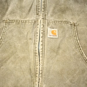 Może przedstawiać: Zbliżenie na brązową kurtkę roboczą Carhartt z zamkiem błyskawicznym i naszywaną kieszenią na lewej piersi. Naszywana kieszeń ma logo Carhartt w kolorze białym.