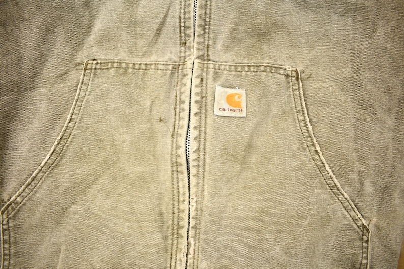 Może przedstawiać: Zbliżenie na brązową kurtkę roboczą Carhartt z zamkiem błyskawicznym i naszywaną kieszenią na lewej piersi. Naszywana kieszeń ma logo Carhartt w kolorze białym.