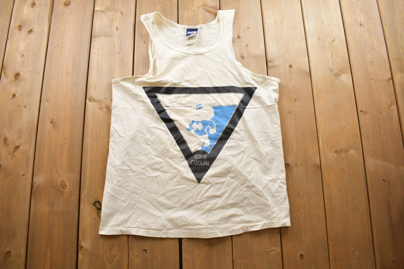 May include: White tank top with a black triangle graphic featuring a blue outline of the state of North Carolina and the text "North Carolina".