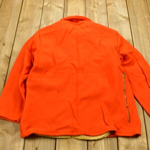 K&ouml;nnte beinhalten: Eine leuchtend orangefarbene Jacke mit einem Rei&szlig;verschluss auf der rechten Seite. Die Jacke ist aus einem dicken, warmen Stoff gefertigt und hat ein braunes Futter.