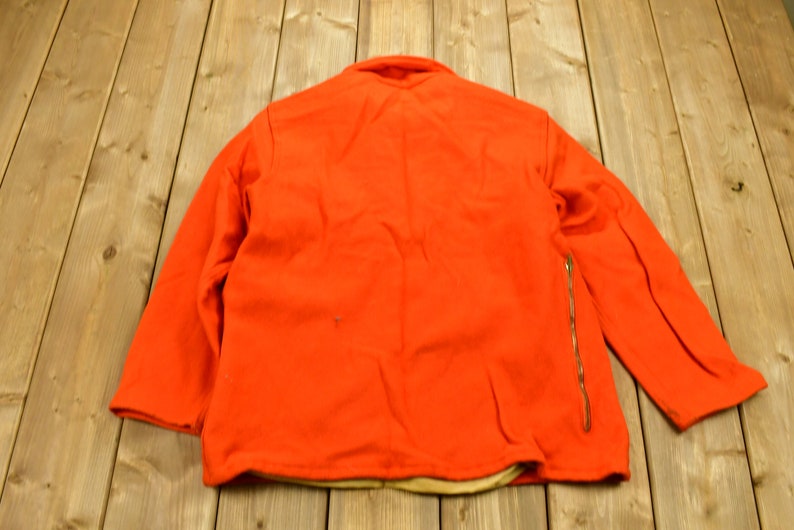 K&ouml;nnte beinhalten: Eine leuchtend orangefarbene Jacke mit einem Rei&szlig;verschluss auf der rechten Seite. Die Jacke ist aus einem dicken, warmen Stoff gefertigt und hat ein braunes Futter.