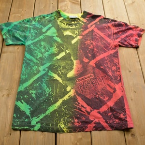 May include: A vintage Reebok Blacktop t-shirt with a tie-dye design in shades of green, yellow, and red. The shirt features a black graphic with the Reebok logo and the text "Blacktop".