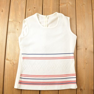 May include: White sleeveless knitted top with red, blue and white stripes at the bottom. The top has a diamond pattern.