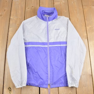 Könnte beinhalten: Eine weiße und violette Windjacke mit Reißverschluss und einem kleinen Logo auf der Brust. Die Jacke hat eine Kapuze mit Kordelzug und elastische Bündchen.