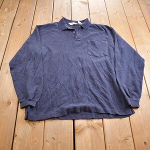 May include: A navy blue long-sleeved polo shirt with a pocket on the left chest. The shirt features a collared neckline and a three-button placket. The fabric appears to be a soft, textured material.