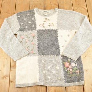 以下が含まれることがあります： グレーとクリーム色のパッチワークセーター。花の刺繍と星のアクセントが施されています。長袖セーターはクルーネックで、テクスチャードニットのデザインが特徴で、居心地の良いヴィンテージ感を演出します。
