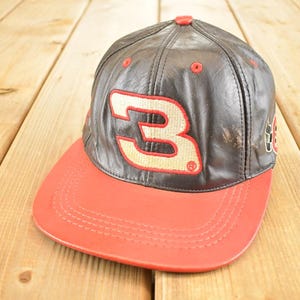 May include: A brown and red baseball cap with the number 3 in a beige and red design. The cap has a curved brim and a button on top. The hat is made of leather-like material.