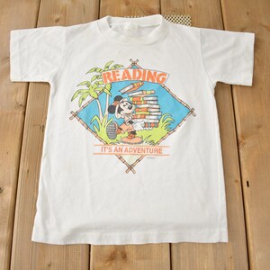 May include: White t-shirt with a cartoon image of Mickey Mouse reading a stack of books. The image is surrounded by a diamond-shaped frame with the words "Reading It's an Adventure".