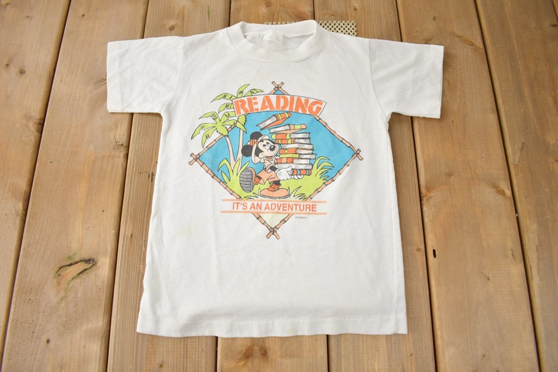 May include: White t-shirt with a cartoon image of Mickey Mouse reading a stack of books. The image is surrounded by a diamond-shaped frame with the words "Reading It's an Adventure".