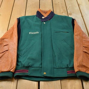 Può includere: Una giacca varsity con corpo in misto lana verde scuro, maniche in pelle color cuoio e colletto blu navy. La parola Wisconsin è ricamata in bianco sul petto destro. Include una chiusura frontale con bottoni a pressione e due tasche laterali.