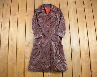 May include: A vintage burgundy trench coat with a button-up front, lapel collar, and matching belt. The coat has a long, flowing design and is made of a shiny, textured material. The interior lining is red.