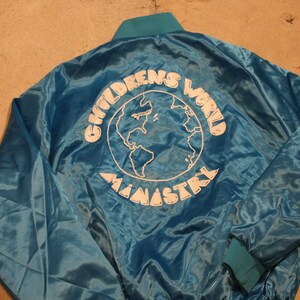 May include: A teal satin jacket with a teal collar and cuffs. The back of the jacket features the words "CHILDREN'S WORLD MINISTRY" in white, surrounding a white outline of the Earth.