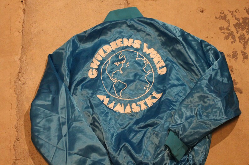 May include: A teal satin jacket with a teal collar and cuffs. The back of the jacket features the words "CHILDREN'S WORLD MINISTRY" in white, surrounding a white outline of the Earth.