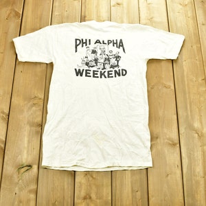 Puede incluir: Camiseta blanca vintage con la inscripción negra "PHI ALPHA WEEKEND" y un dibujo de personas. La camiseta está colocada sobre una superficie de madera.