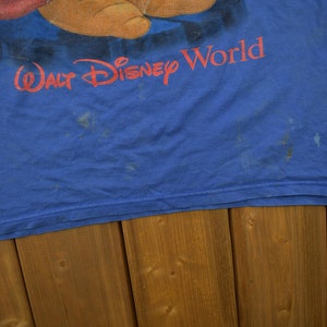 Pu&ograve; includere: Una maglietta blu con un'immagine sbiadita di un personaggio dei cartoni animati e il testo "Walt Disney World".
