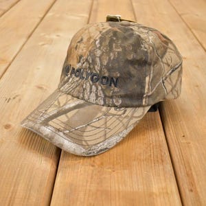 May include: Camouflage baseball cap with the word "POLYGON" embroidered in black. The cap features a curved brim and an adjustable strap. The hat is made of a durable fabric and is designed for outdoor activities.