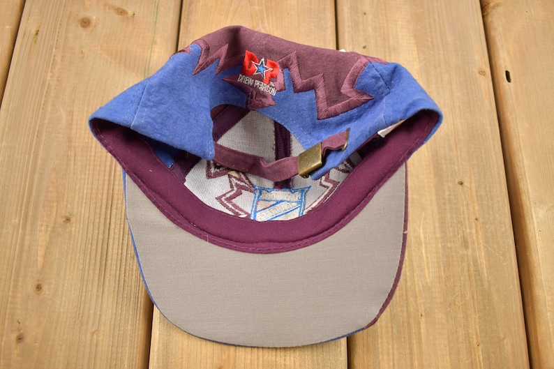 May include: A blue and purple baseball cap with a white and blue embroidered logo that says "D.P. Drew Pearson" and a red star. The cap has a gray bill.