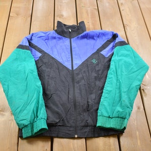 Puede incluir: Una chaqueta cortavientos negra, azul y verde con cierre de cremallera. La chaqueta tiene un pequeño logotipo en el pecho izquierdo.