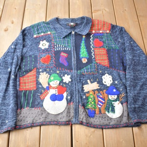 Könnte beinhalten: Ein blauer Strickcardigan mit Patchwork-Design, das Schneemänner, einen Weihnachtsbaum und andere festliche Motive zeigt. Der Cardigan hat einen Reißverschluss und einen Kragen.