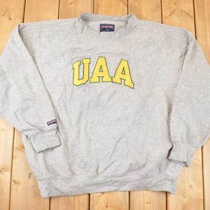 May include: A heather gray sweatshirt with the word "UAA" in yellow block letters across the chest. The sweatshirt has a crew neck, long sleeves, and ribbed cuffs. A small tag is visible on the collar.