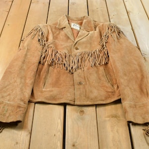 May include: A tan suede jacket featuring long fringe detailing across the chest, shoulders, and sleeves. The jacket has a collared neckline, a button-front closure, and two vertical slit pockets. A white brand label is visible inside the collar.