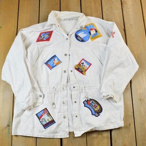 May include: White jacket with multiple colorful embroidered patches. The patches feature various designs, including nautical themes and cartoon characters. The jacket has a button-up front, a drawstring waist, and buttoned cuffs. The jacket is laid flat on a wooden surface.