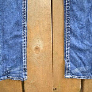 K&ouml;nnte beinhalten: Eine blaue Jeans aus Denim mit wei&szlig;en N&auml;hten entlang der N&auml;hte. Die Jeans h&auml;ngt an einer Holzoberfl&auml;che.