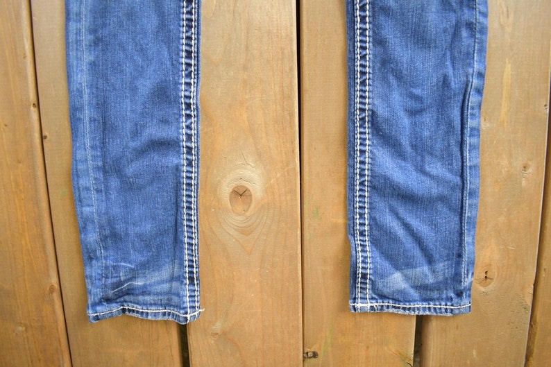 K&ouml;nnte beinhalten: Eine blaue Jeans aus Denim mit wei&szlig;en N&auml;hten entlang der N&auml;hte. Die Jeans h&auml;ngt an einer Holzoberfl&auml;che.