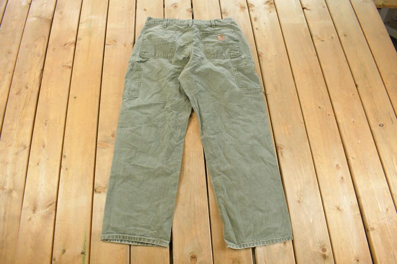 Puede incluir: Vista trasera de pantalones de trabajo verde oliva con m&uacute;ltiples bolsillos y un peque&ntilde;o logotipo. Los pantalones est&aacute;n hechos de una tela duradera y tienen un ajuste relajado, adecuados para diversas tareas.