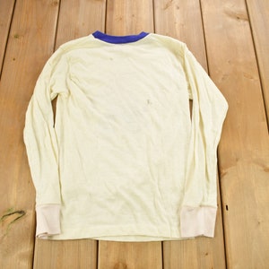 May include: A long-sleeved, white t-shirt with a blue trim around the neckline. The shirt is made of a soft, lightweight fabric and has a relaxed fit.
