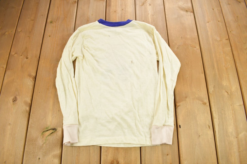 May include: A long-sleeved, white t-shirt with a blue trim around the neckline. The shirt is made of a soft, lightweight fabric and has a relaxed fit.