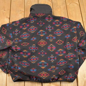 May include: Black fleece jacket featuring a geometric pattern in red, blue, and orange. The jacket has a high collar and a full zip. The pattern is repeated across the entire jacket, creating a striking design.
