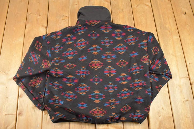 May include: Black fleece jacket featuring a geometric pattern in red, blue, and orange. The jacket has a high collar and a full zip. The pattern is repeated across the entire jacket, creating a striking design.