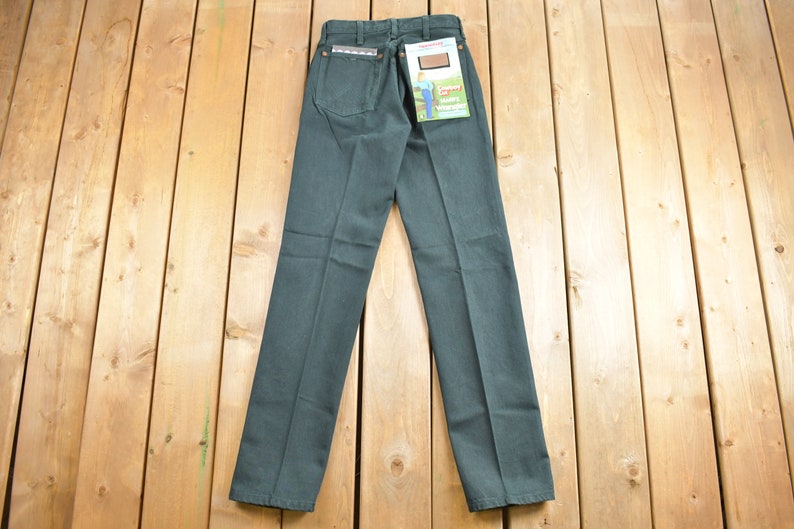 Op de afbeelding: Een donkergroene jeans met een bruine leren patch op de achterzak. De jeans heeft een rechte pijp en een klassieke pasvorm. De jeans ligt plat op een houten oppervlak.