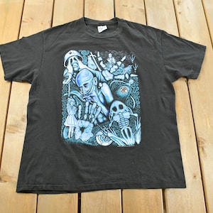 May include: Dark grey t-shirt with a surreal blue and white graphic. The design features figures, a skeleton, and abstract elements. The t-shirt is displayed on a wooden surface.