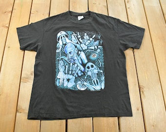 Puede incluir: Camiseta gris oscuro con un gráfico surrealista en tonos azules y blancos. El diseño incluye figuras, un esqueleto y elementos abstractos. La camiseta está colocada sobre una superficie de madera.