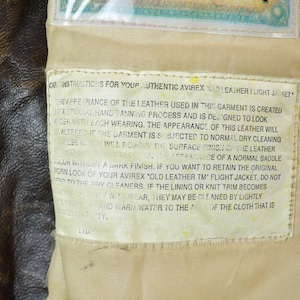 May include: Close-up of a brown leather jacket with a zipper and a tag with care instructions. The tag reads "Authentic Avirex Old Leather Flight Jacket". The leather has a worn appearance.